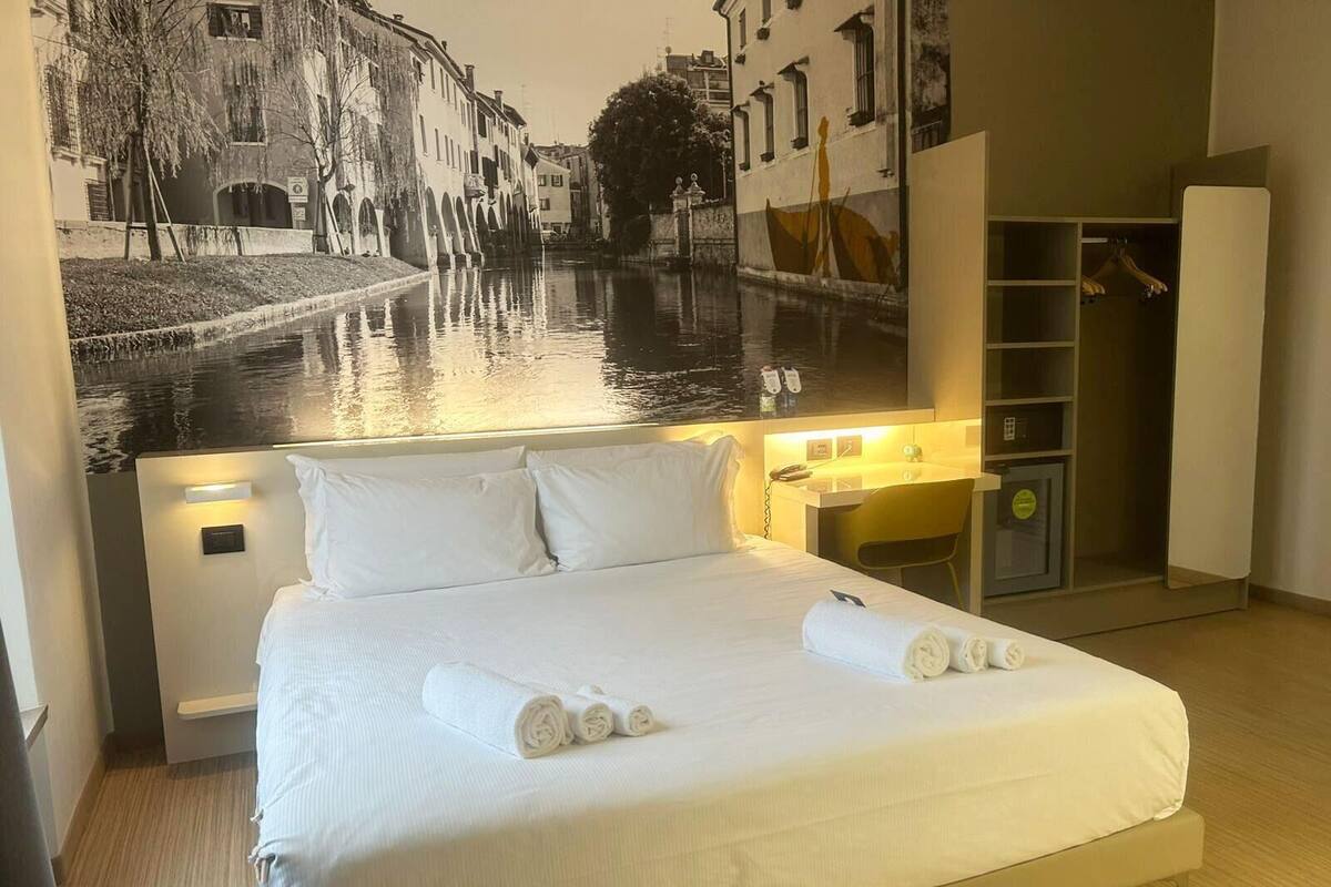 A hotel room with a bed, a nightstand, a mirror, a wardrobe, and a large wall mural of a canal scene.