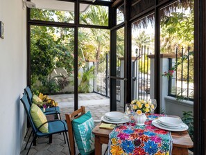 A covered patio with a table set for dining, chairs, and a view of a garden. - Cozy studio with all the extras. Fully equipped kitchen, TV, free wifi, pool. (Puerto Morelos)