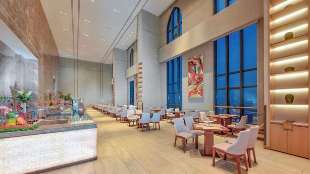 A spacious hotel lobby with a long reception desk, multiple seating areas, and large windows offering views of the cityscape.