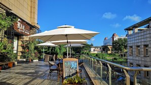 An outdoor café with wooden tables and white umbrellas, overlooking a river and surrounded by greenery and buildings.