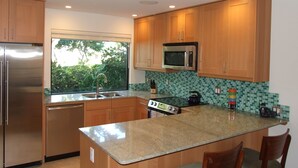 A modern kitchen with wooden cabinets, stainless steel appliances, a large window, and a green-tiled backsplash. - Ekahi 36a, 1BR 2BA condo, Sleeps 4 (2 Adults + 2 Kids) (Wailea-Makena)