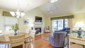 A spacious living room with a fireplace, a dining area with a wooden table and chairs, a ceiling fan, and large windows allowing natural light.