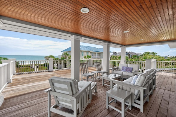 A covered outdoor deck with white furniture, a view of the ocean, and a house in the background.