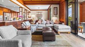 A spacious living area with a large sectional sofa, a coffee table, and a fireplace. There is a kitchen area with wooden cabinets and a refrigerator. The room has a high ceiling with wooden beams and large windows. - Top-Floor breathtaking views, close to beach! (Kamuela)