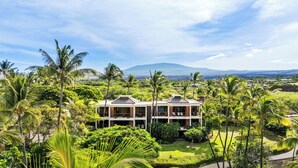 A modern two-story building surrounded by lush greenery and palm trees, with a view of distant mountains. - Top-Floor breathtaking views, close to beach! (Kamuela)