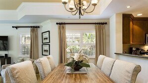 A dining area with a wooden table, beige upholstered chairs, a chandelier, and a window with curtains. - Ground Floor Luxury Condo | Fully Furnished | Very Close to Disney! (Kissimmee)
