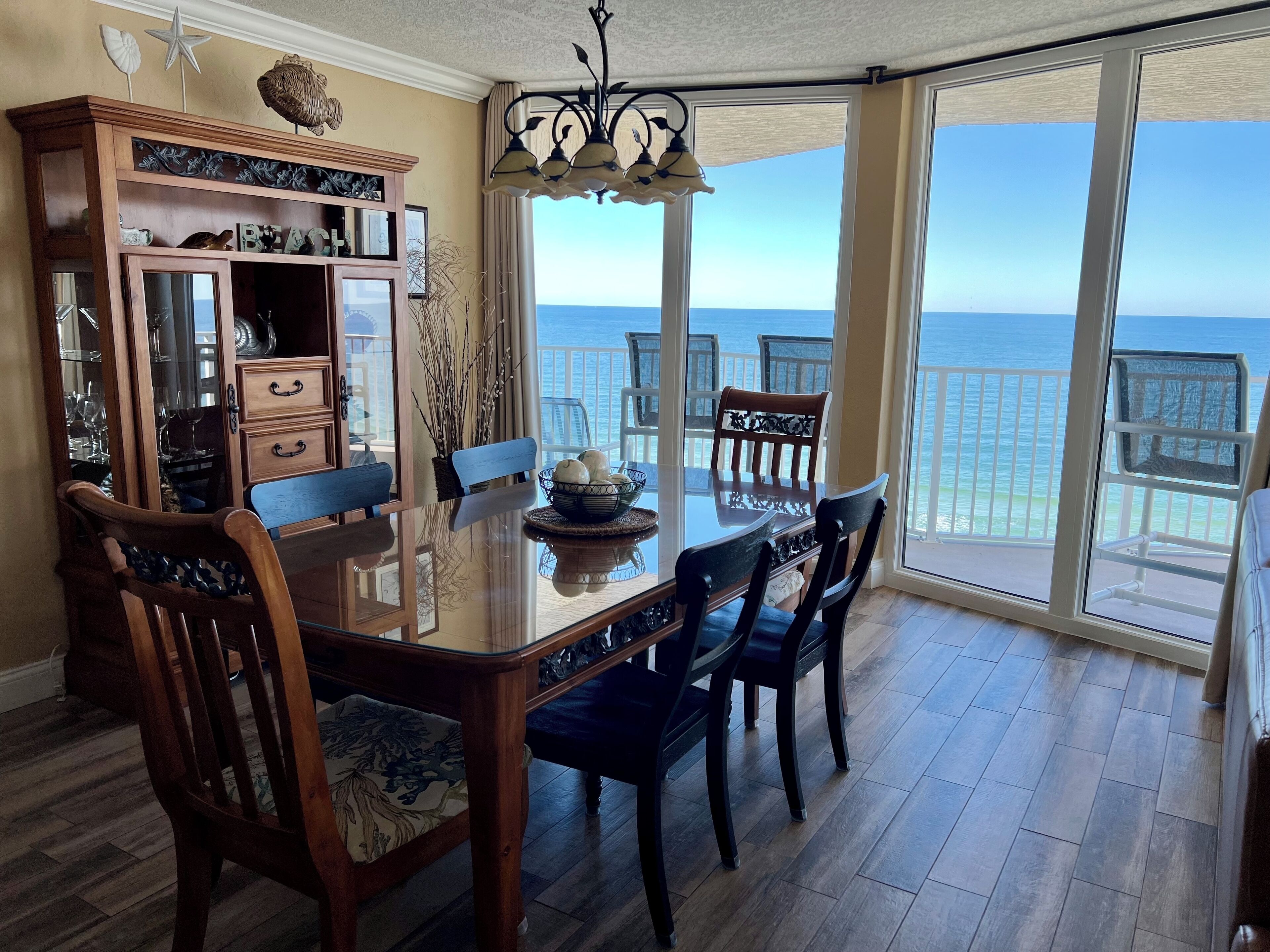 A dining area with a glass table and chairs, a wooden cabinet with glass doors, and a view of the ocean through large windows.