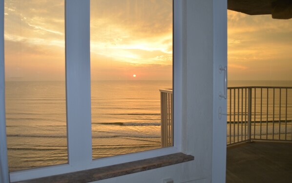 A view through a window of a sunset over the ocean, with a railing and a door visible. - Updated Direct Oceanfront - 3 bedroom #1007; Beach Access and Heated Pool (Daytona Beach Shores)