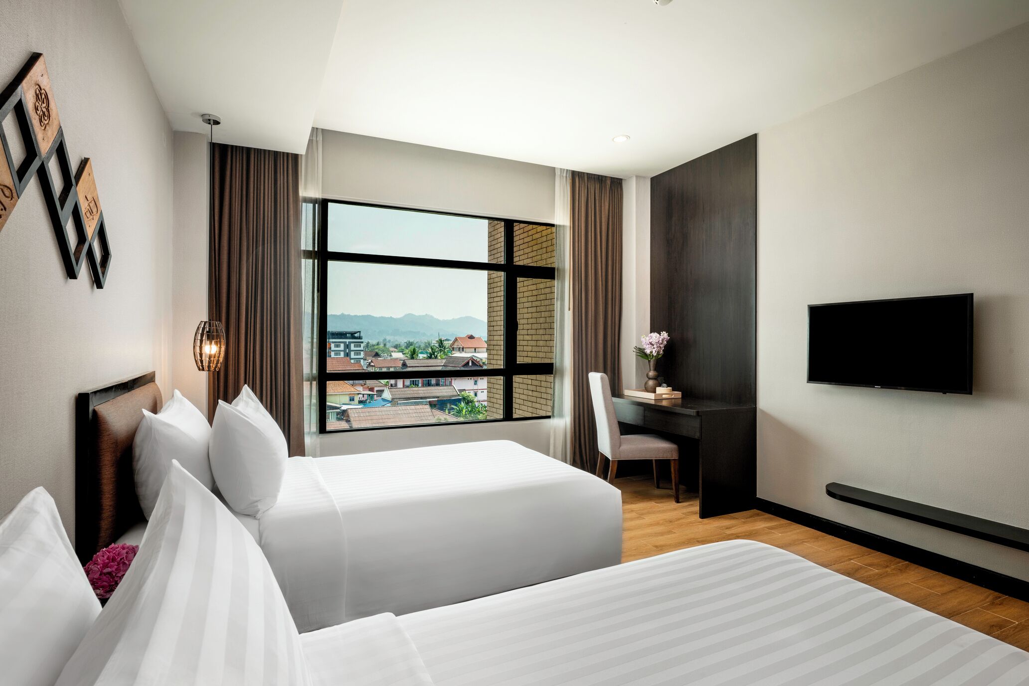 Habitació d'hotel amb dos llits, una taula, una cadira, un televisor i una finestra gran amb vista a edificis i muntanyes.