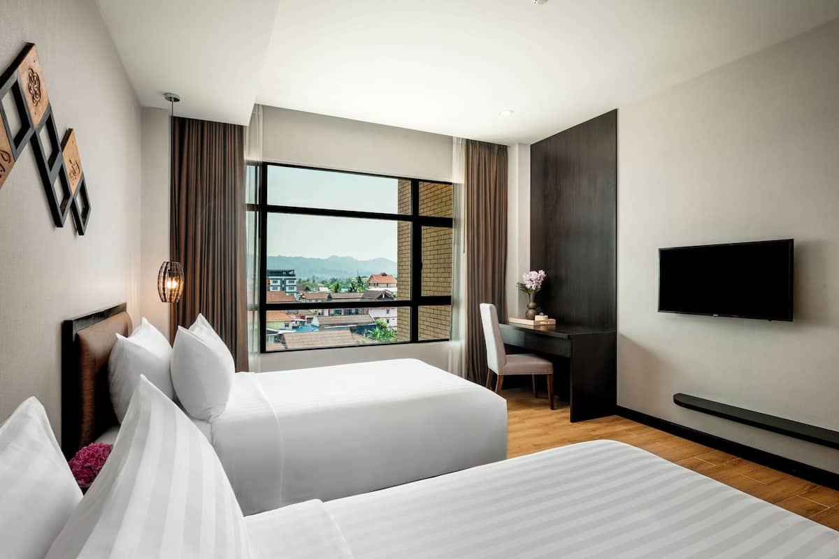 Ein Hotelzimmer mit zwei Betten, einem Schreibtisch, einem Stuhl, einem Fernseher und einem großen Fenster mit Blick auf Gebäude und Berge.