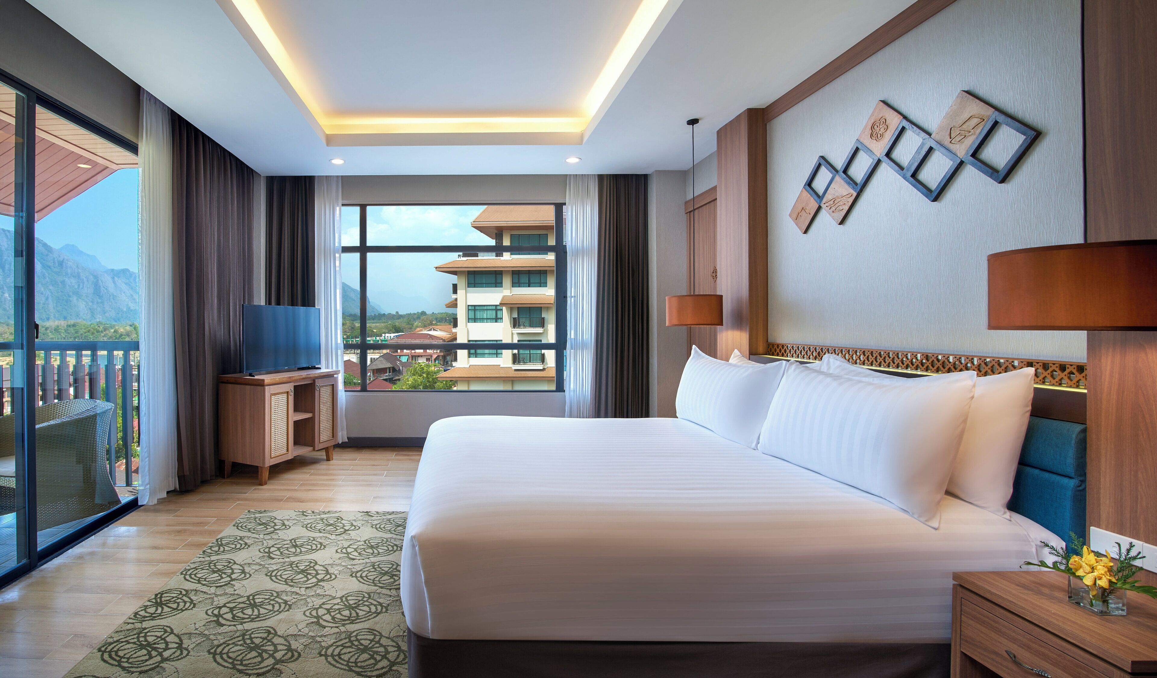 A hotel room with a large bed, a TV on a wooden stand, a nightstand with a lamp, and a view of buildings and mountains through the window.