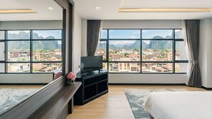 Habitación de hotel con una cama grande, un televisor sobre un mueble de madera, un espejo y vistas de un paisaje urbano a través de amplios ventanales.