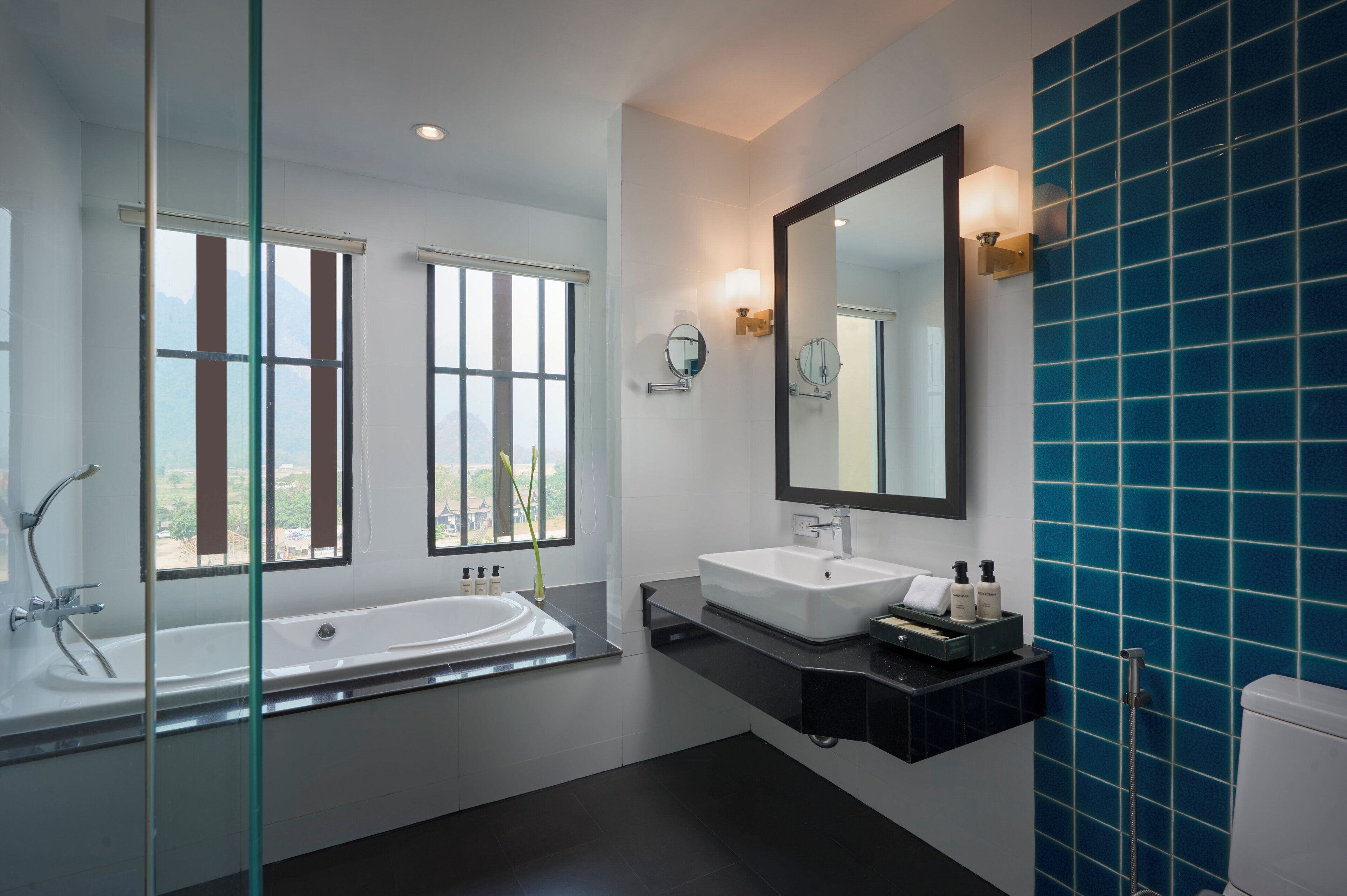 A modern bathroom with a bathtub, two sinks, a large mirror, and a view of the outdoors through large windows.