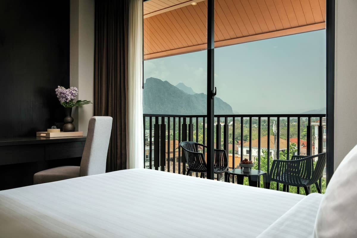 A hotel room with a large bed, a desk with a vase of flowers, a balcony with chairs and a table, and a view of mountains.