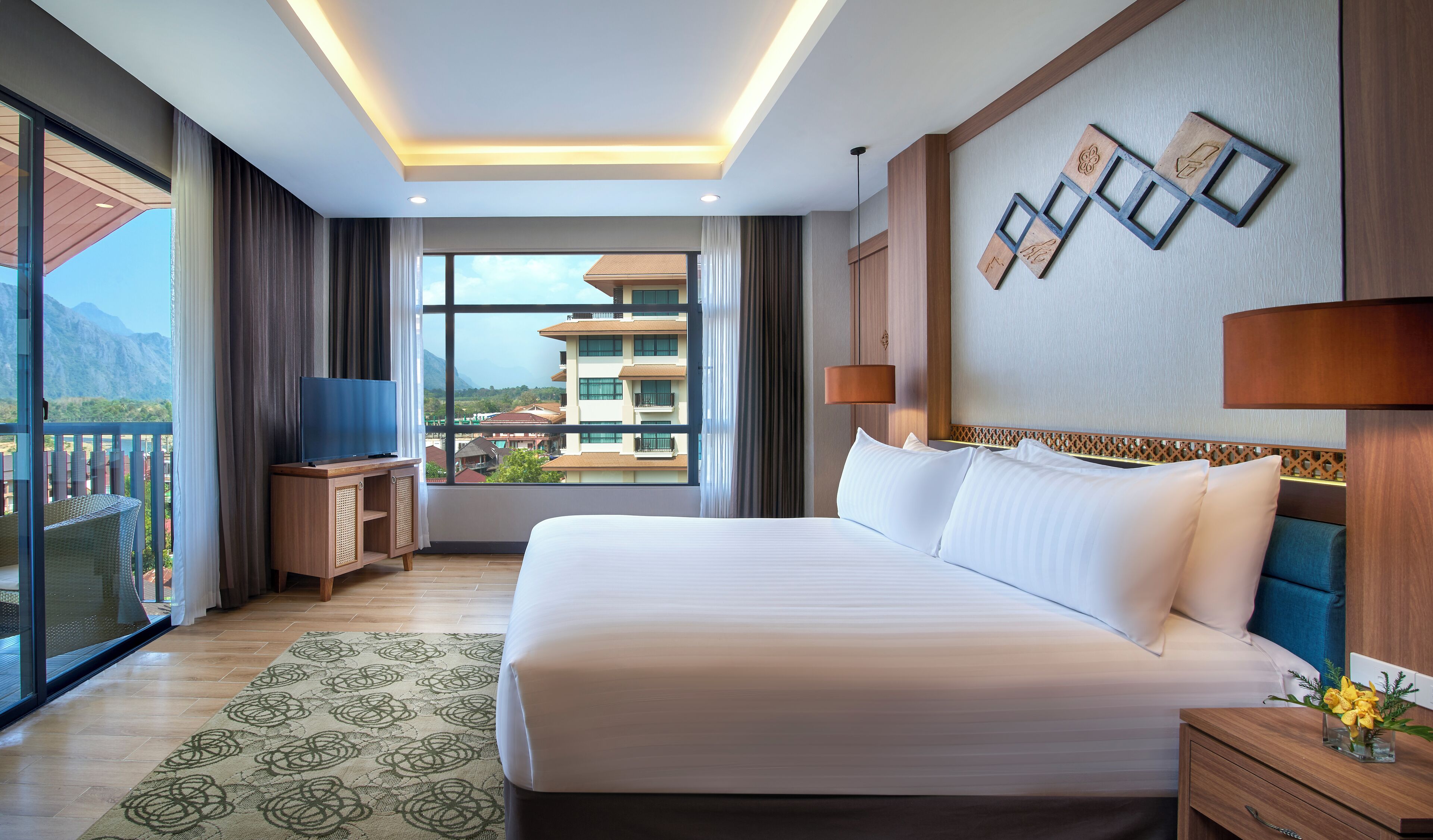 Habitación de hotel con una cama grande, un televisor sobre un soporte de madera, vistas a edificios y montañas a través de la ventana, y un balcón con una silla de mimbre.