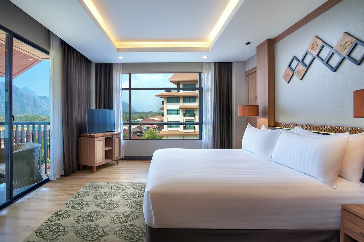A hotel room with a large bed, a TV on a wooden stand, a view of buildings and mountains through the window, and a balcony with a wicker chair.