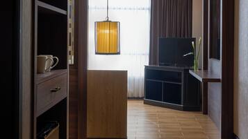 Habitación de hotel moderna con suelo de madera, televisor sobre un mueble de madera, pequeña área de cocina y un ventanal con cortinas.
