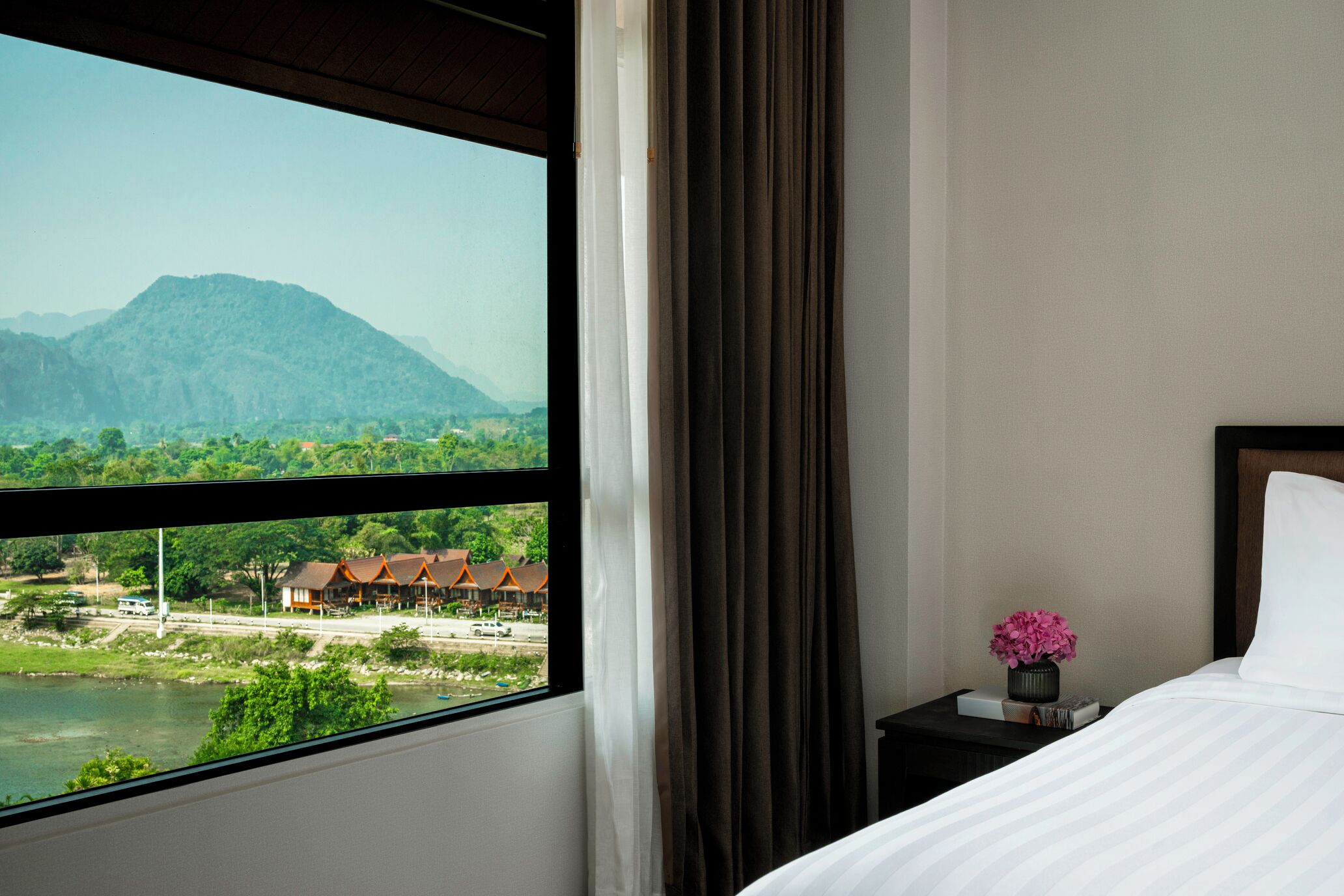A hotel room with a large window, a bed, a nightstand with a vase of flowers, and a view of a mountain and buildings.