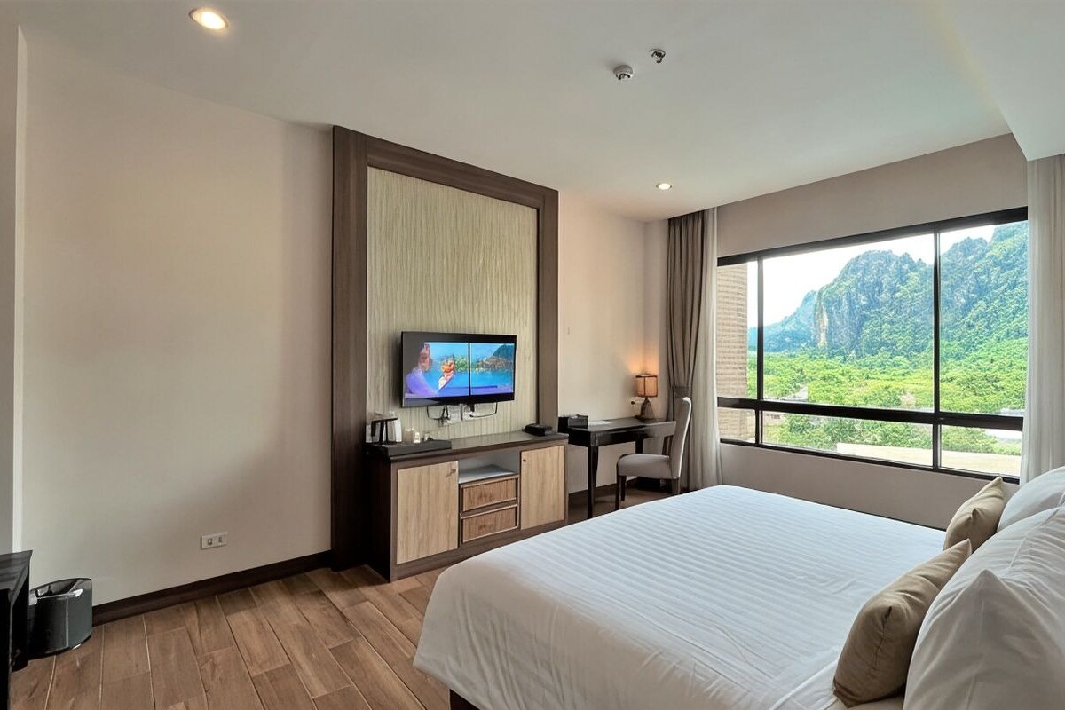 A hotel room with a large bed, a desk with a chair, a television, and a window with a view of greenery.