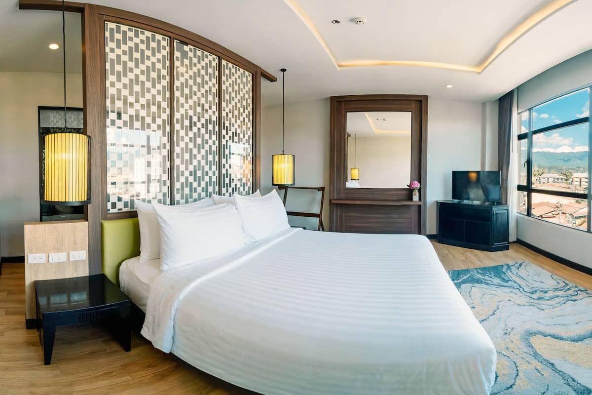 Ein modernes Hotelzimmer mit einem großen Bett, einem Schreibtisch, einem Fernseher und Blick auf die Stadt durch große Fenster.