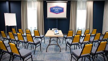 Un salón de conferencias con una pantalla de presentación que muestra 'Hampton Inn by Hilton', una mesa con una computadora portátil y filas de sillas.