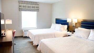 A hotel room with two beds, a desk with a lamp, a chair, and a window with a plaid valance.