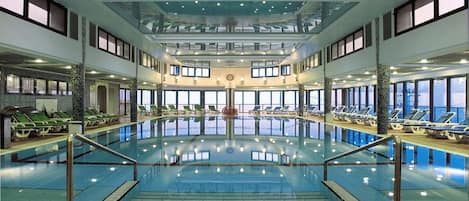 A large indoor swimming pool with multiple levels, sunlit windows, and lounge areas.