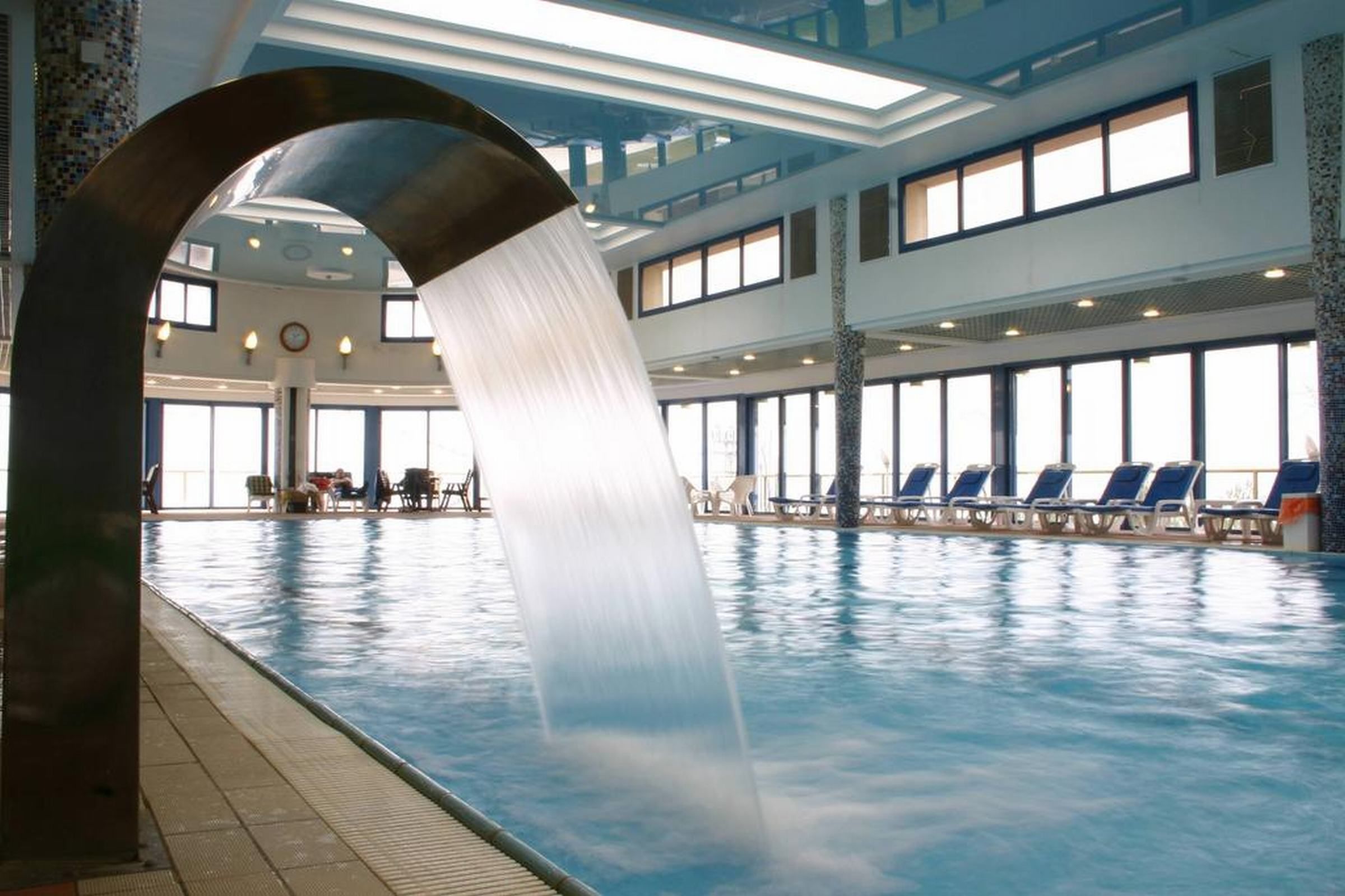 Una piscina interior con fuente de agua, sillas de descanso y un techo alto con tragaluces.