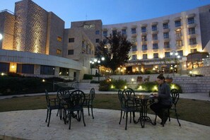 Un área exterior con mesas y sillas, un edificio con varias ventanas y un árbol en primer plano.