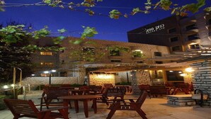 An outdoor patio area with wooden tables and chairs, stone walls, and a building with a sign in the background. - Canaan Spa Hotel (Safed)