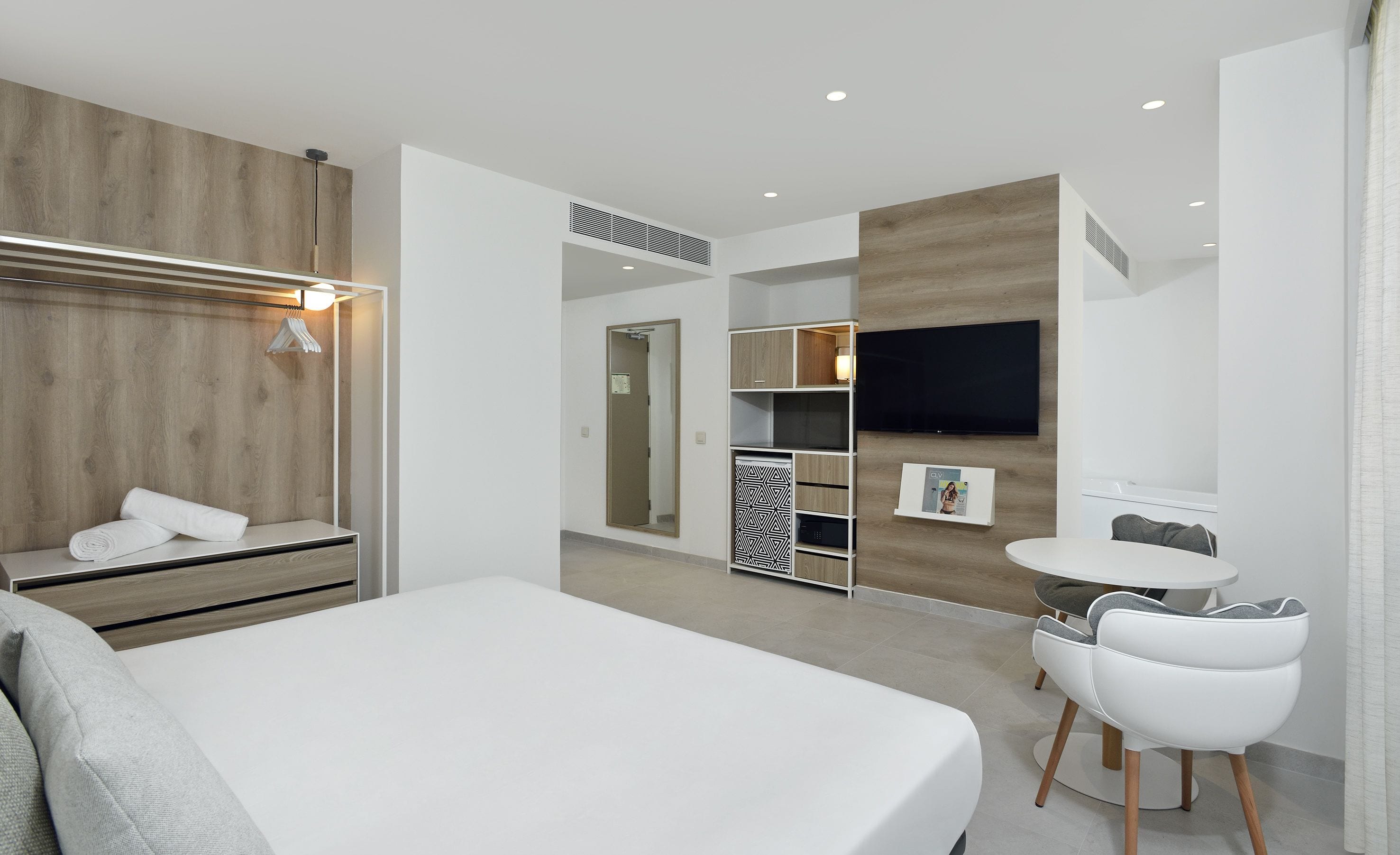 Una habitación de hotel moderna con una cama grande, un armario con espejo, una zona de estar con mesa y sillas, y un televisor montado en la pared.