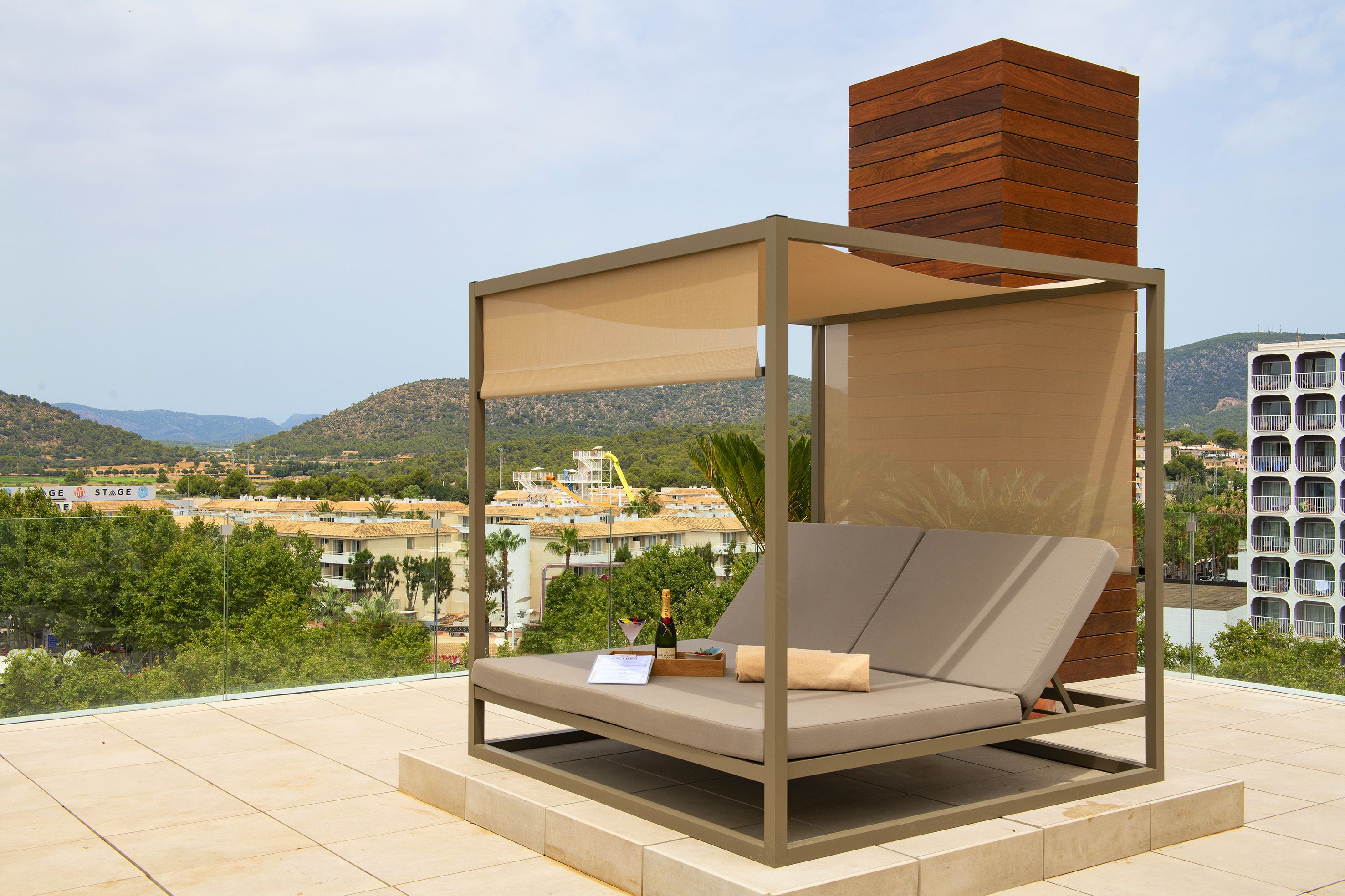 A sun lounger with a canopy, a bottle of champagne, and a menu on a terrace with a city view.