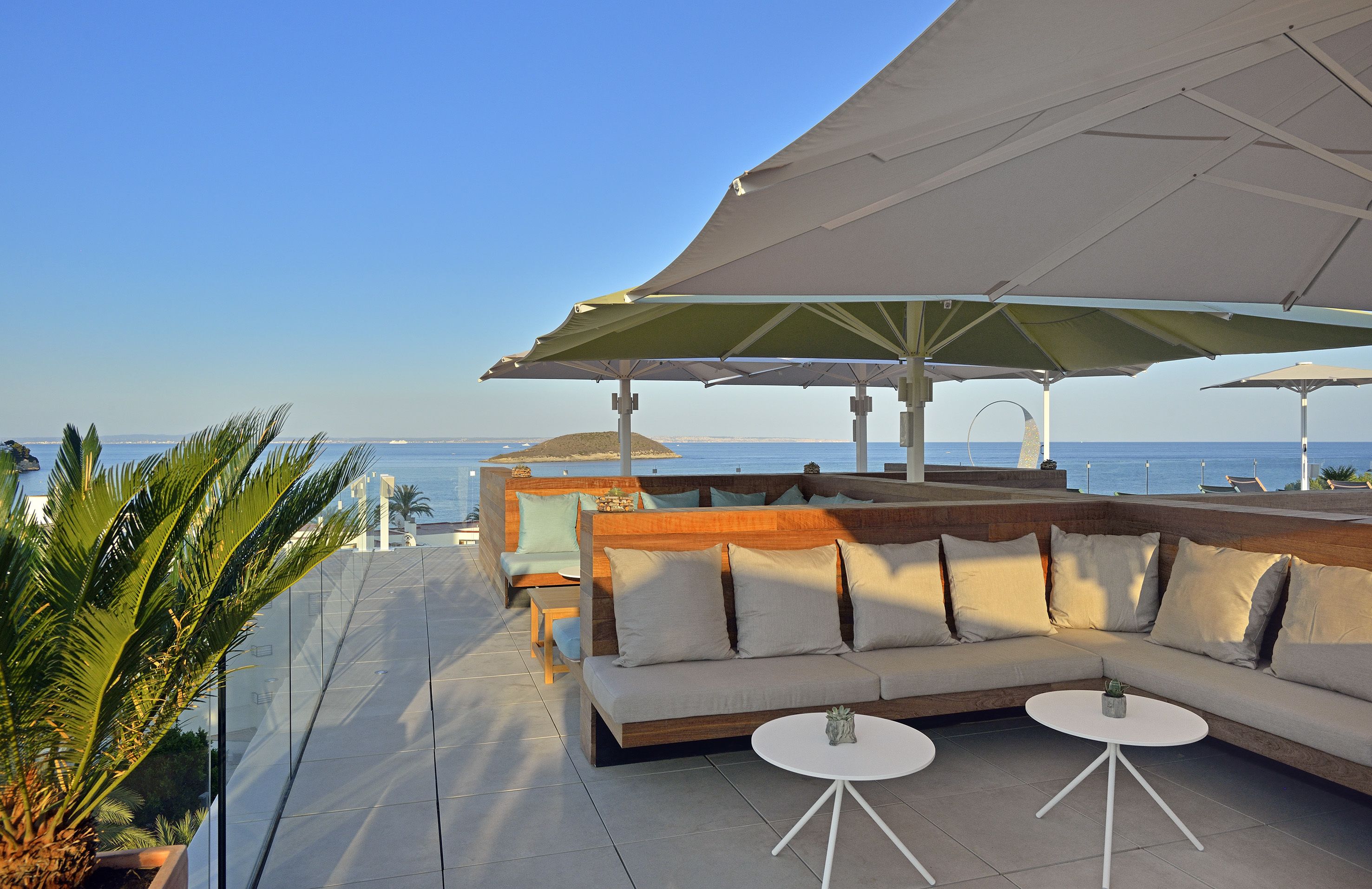 A rooftop terrace with a large sectional sofa, two small round tables, and a large umbrella providing shade.