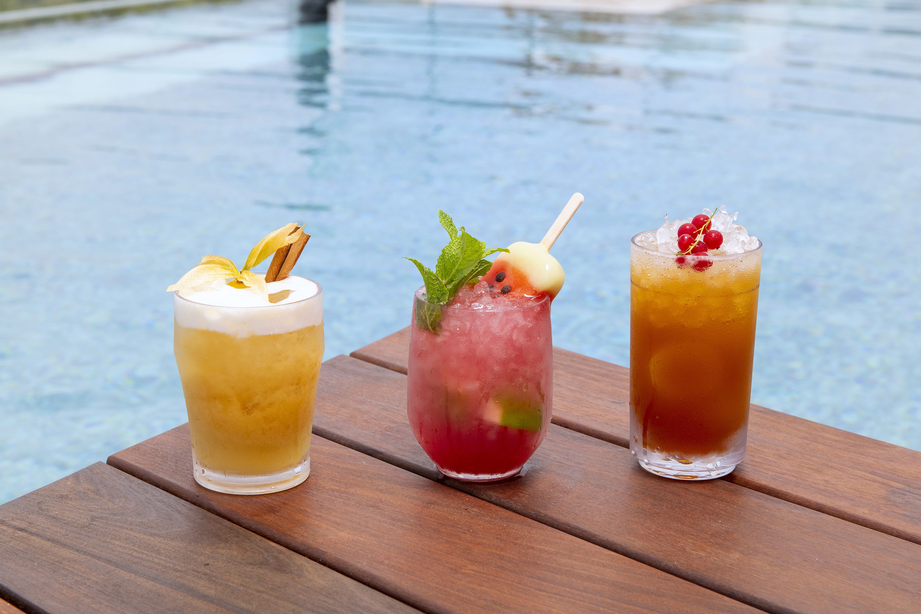Three drinks on a wooden table by a pool.