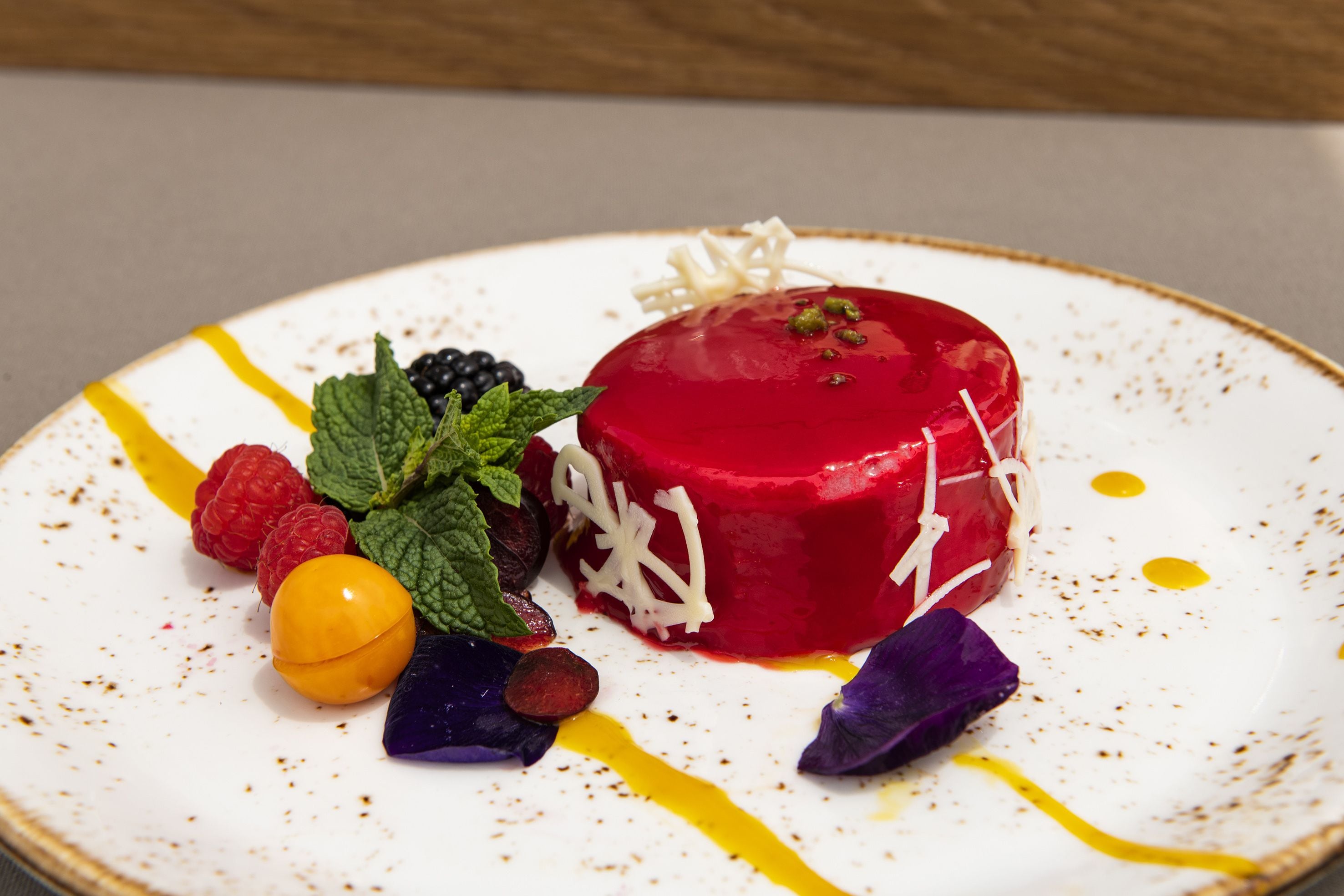 Un dessert con un cuore rosso, circondato da diversi frutti e foglie di menta su un piatto bianco con macchie gialle e marroni.