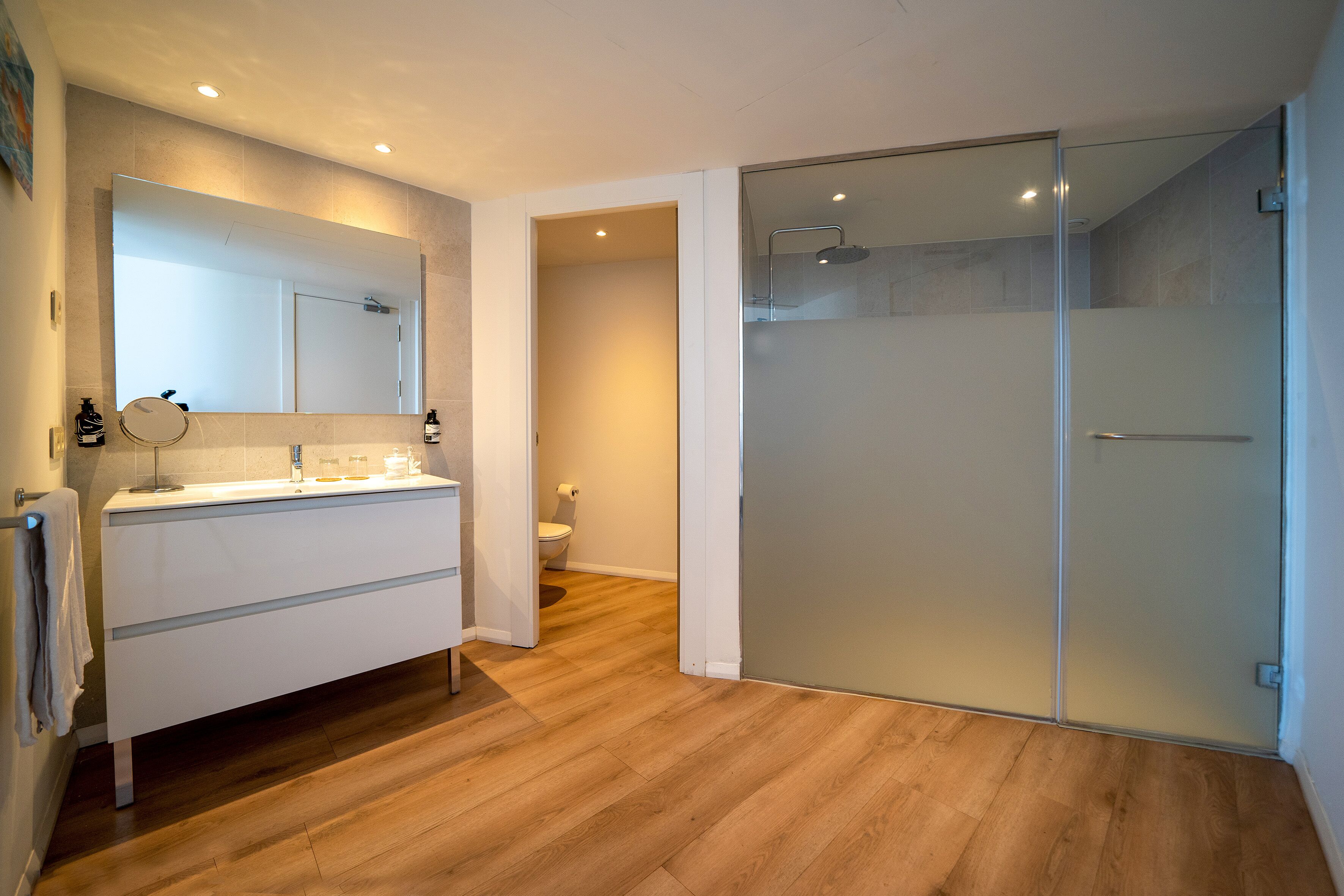 A modern bathroom with a large mirror, a glass shower enclosure, and a wooden floor.
