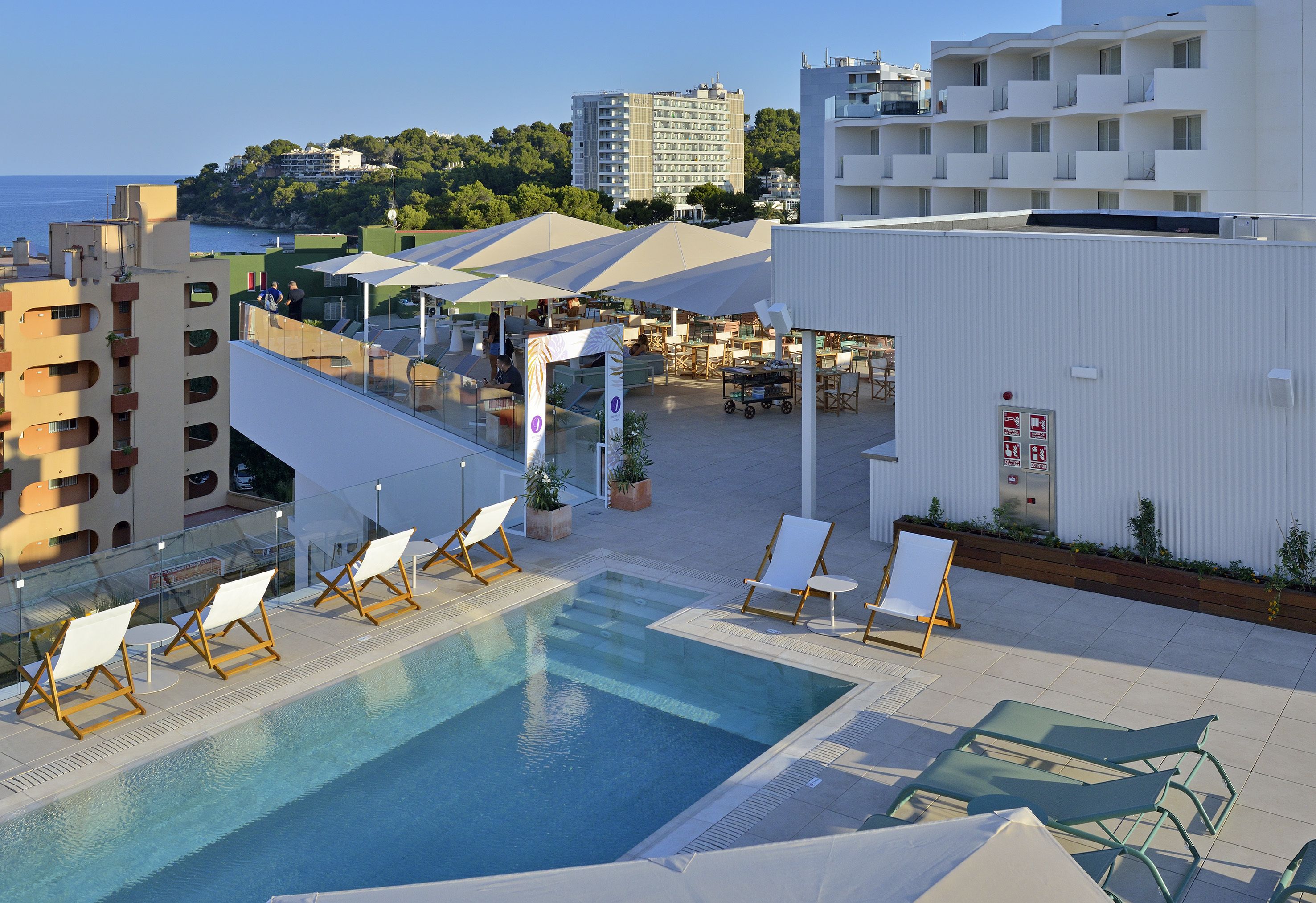 A rooftop pool area with lounge chairs, umbrellas, and a view of the sea and buildings.