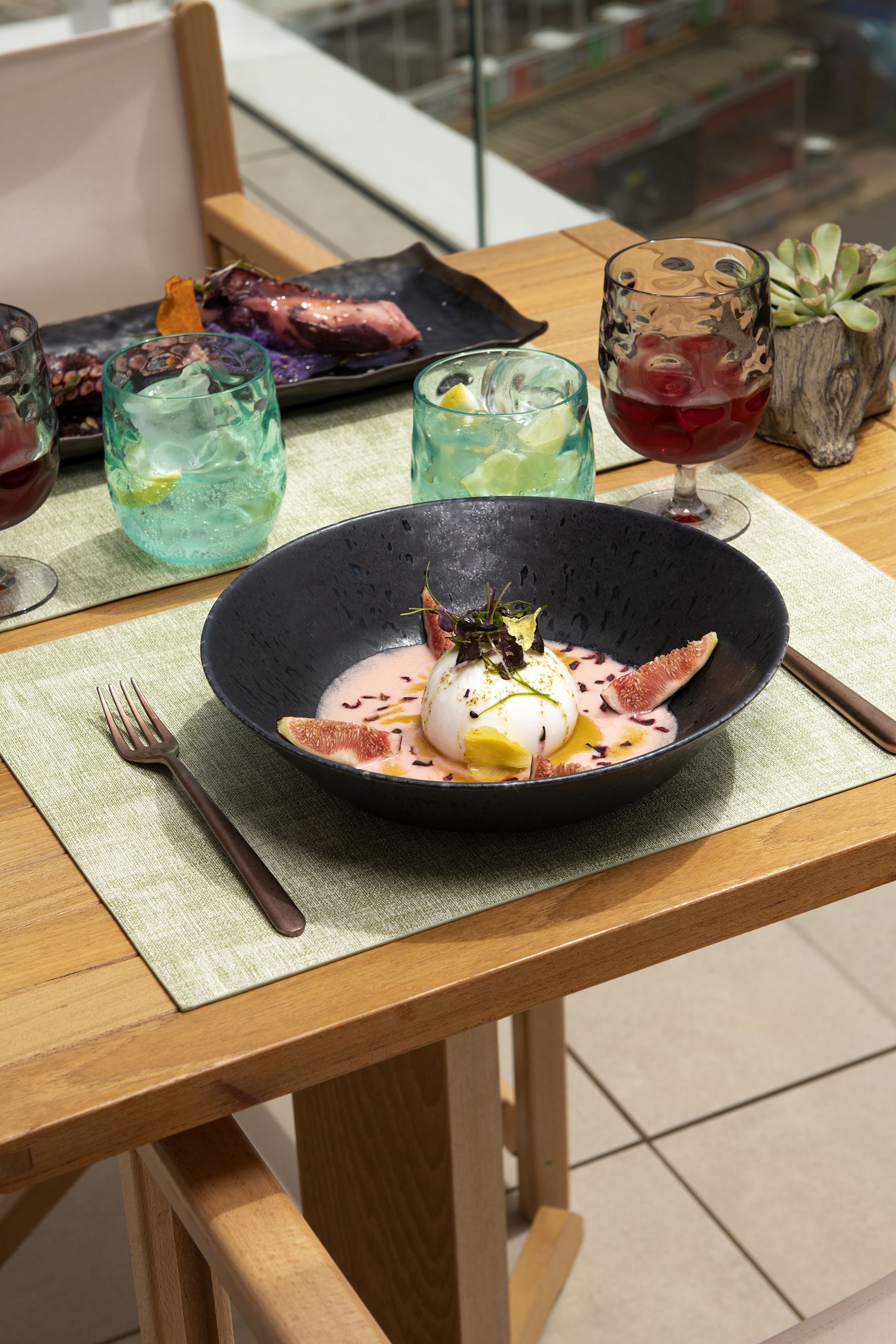 Une table à manger avec un bol noir contenant un plat, deux verres remplis de boissons et une assiette avec de la nourriture.