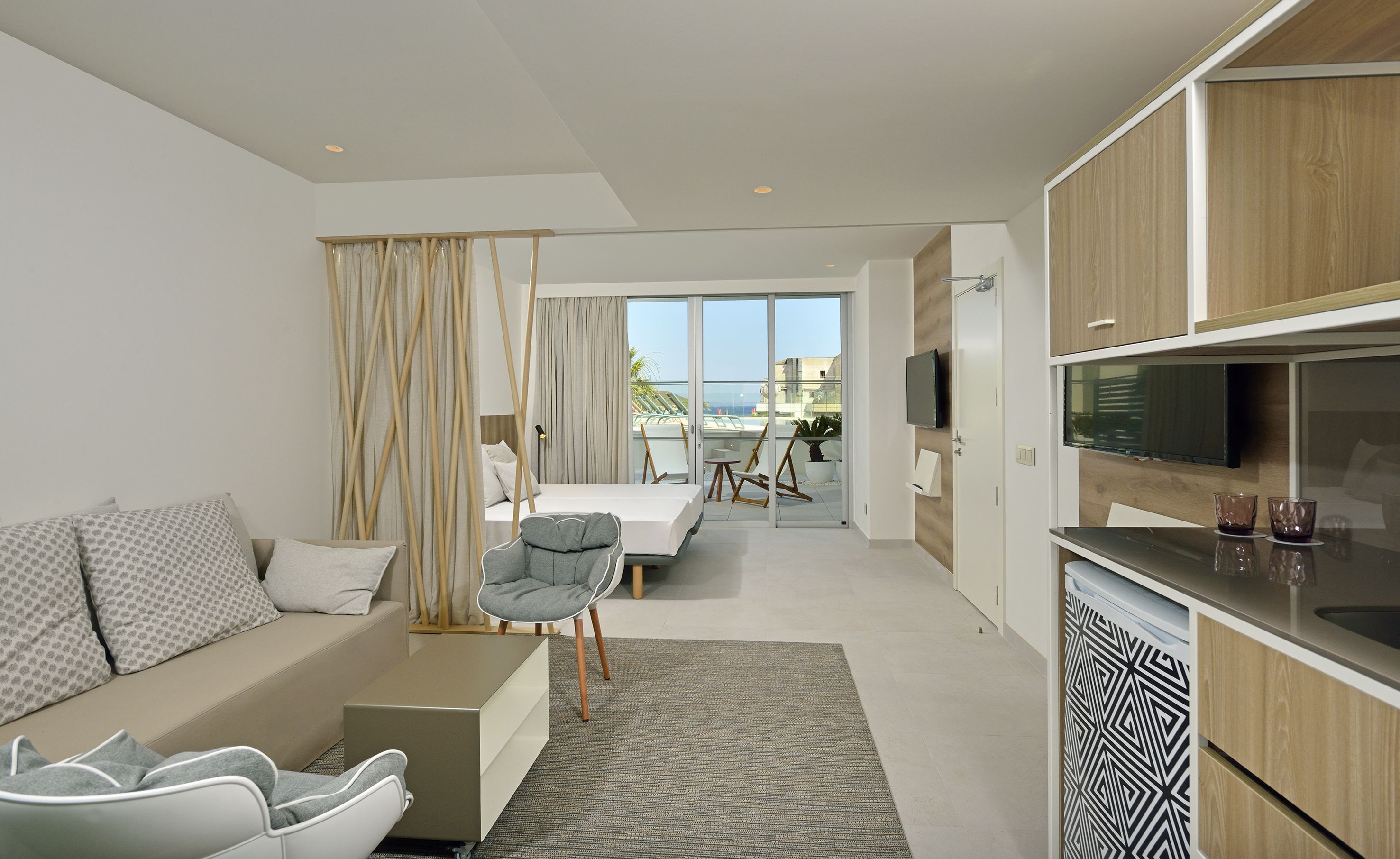 Une chambre d’hôtel moderne dotée d’un canapé, d’une petite table à manger, d’un coin cuisine et d’un balcon offrant une vue sur la mer.