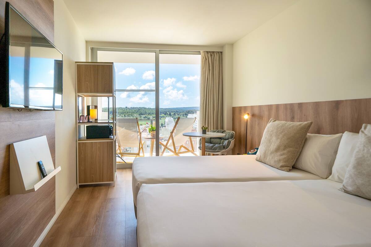 Une chambre d’hôtel avec deux lits, une télévision, une petite table avec une lampe et un balcon offrant une vue sur la ville.