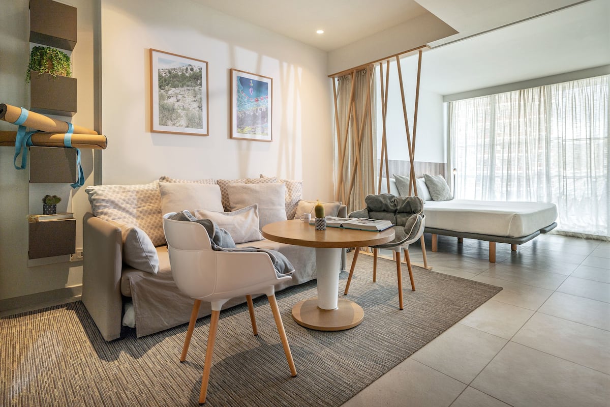 Un salon moderne avec un canapé, une table basse ronde et un coin repas avec des chaises. Deux tableaux encadrés ornent le mur, une plante est posée sur une étagère et une fenêtre est agrémentée de rideaux.