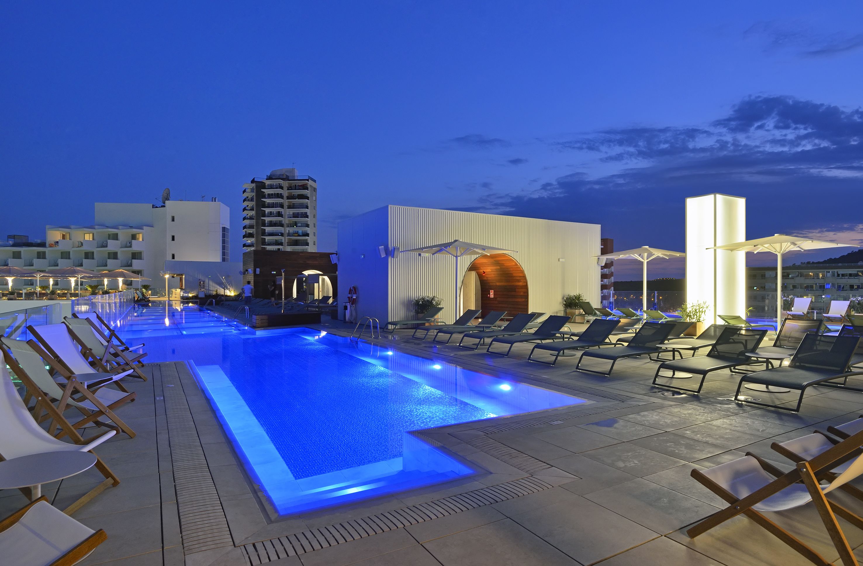 Zona de piscina en la azotea con sillones de descanso y sombrillas, rodeada de edificios modernos.