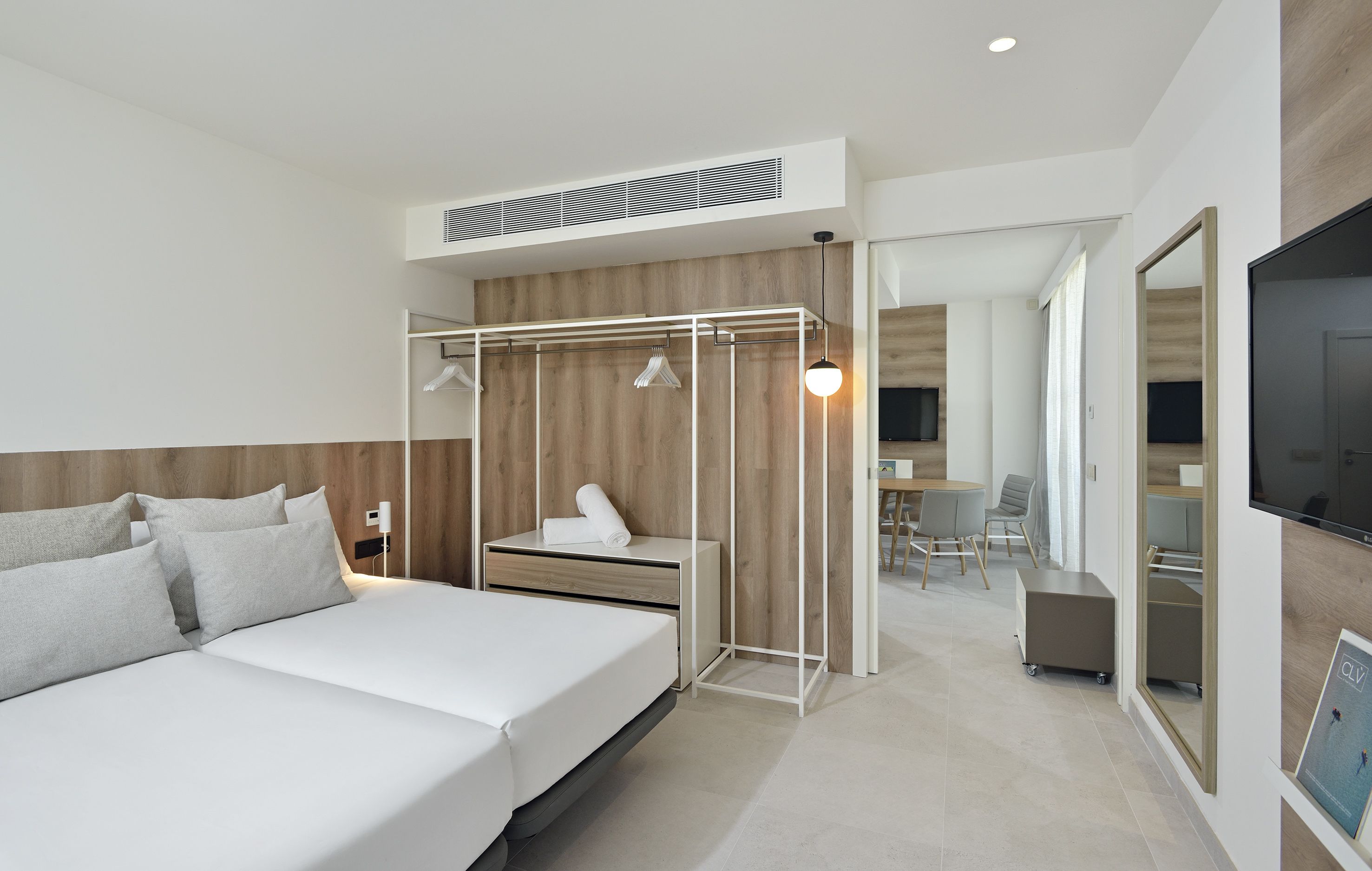 A modern hotel room with a large bed, a wooden headboard, a desk with a chair, a TV, and a wardrobe with a mirror.