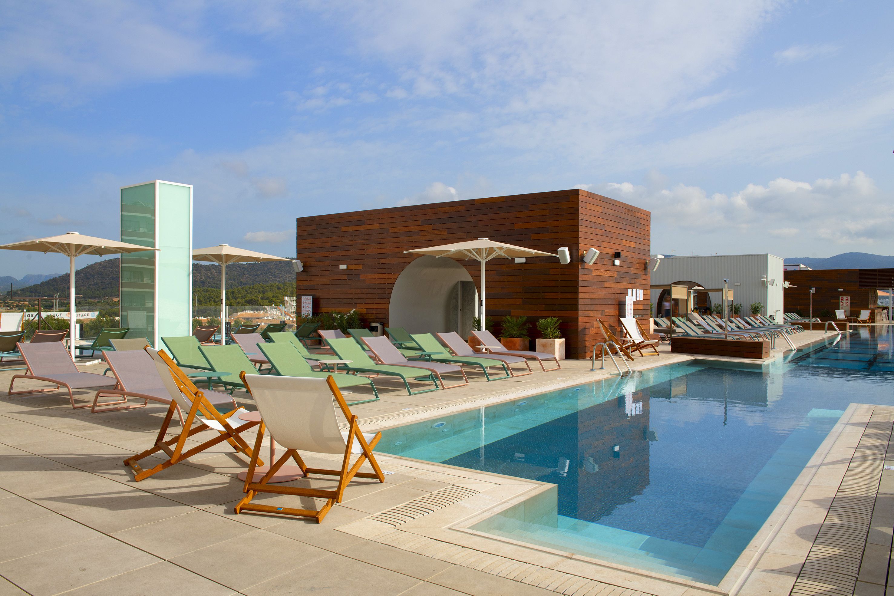 Un área junto a la piscina con sillones de descanso y sombrillas, un edificio moderno con fachada de madera, y un cielo azul despejado.