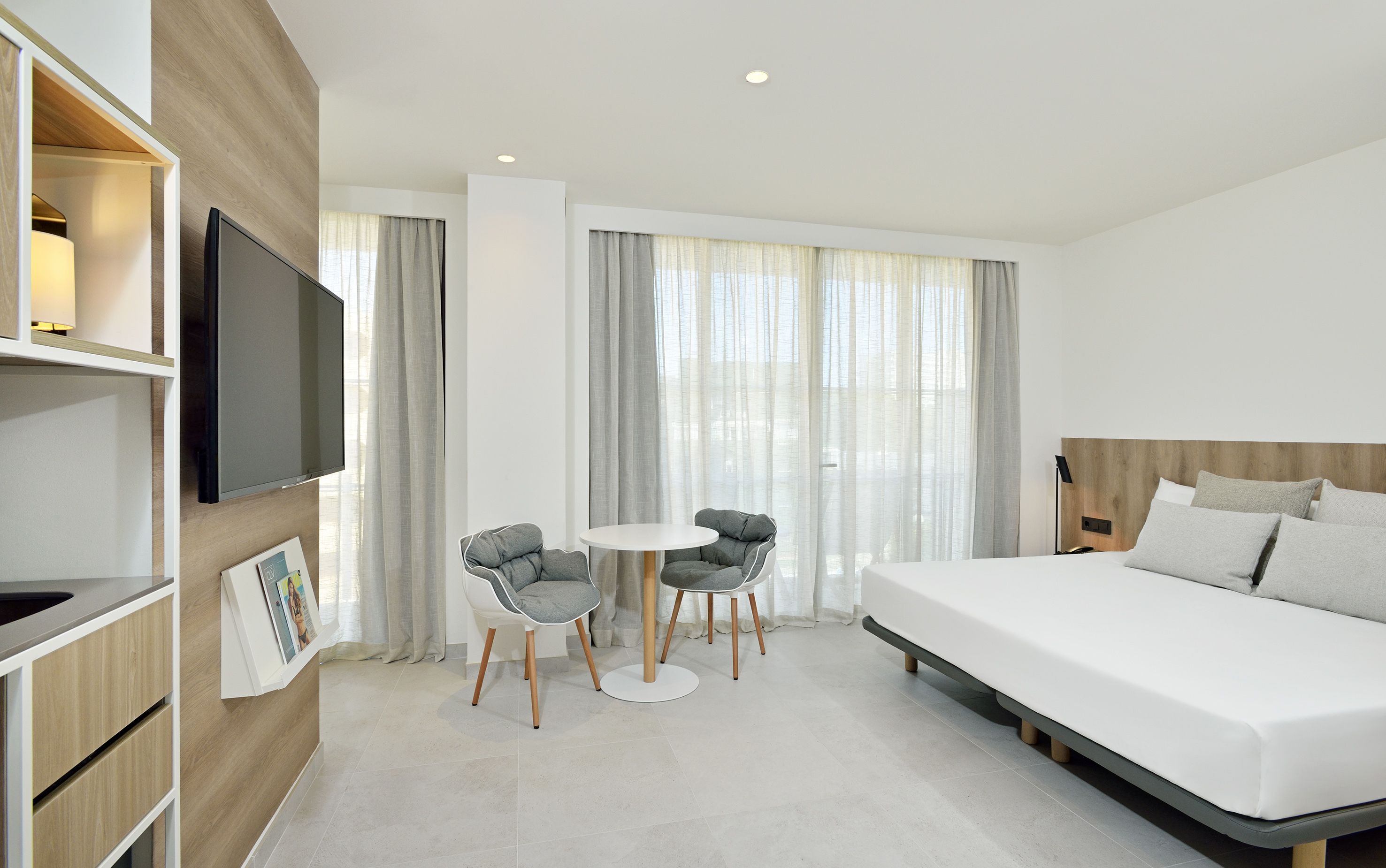 Une chambre d’hôtel moderne avec un grand lit, un téléviseur fixé au mur, une petite table à manger avec deux chaises et une grande fenêtre avec des rideaux.