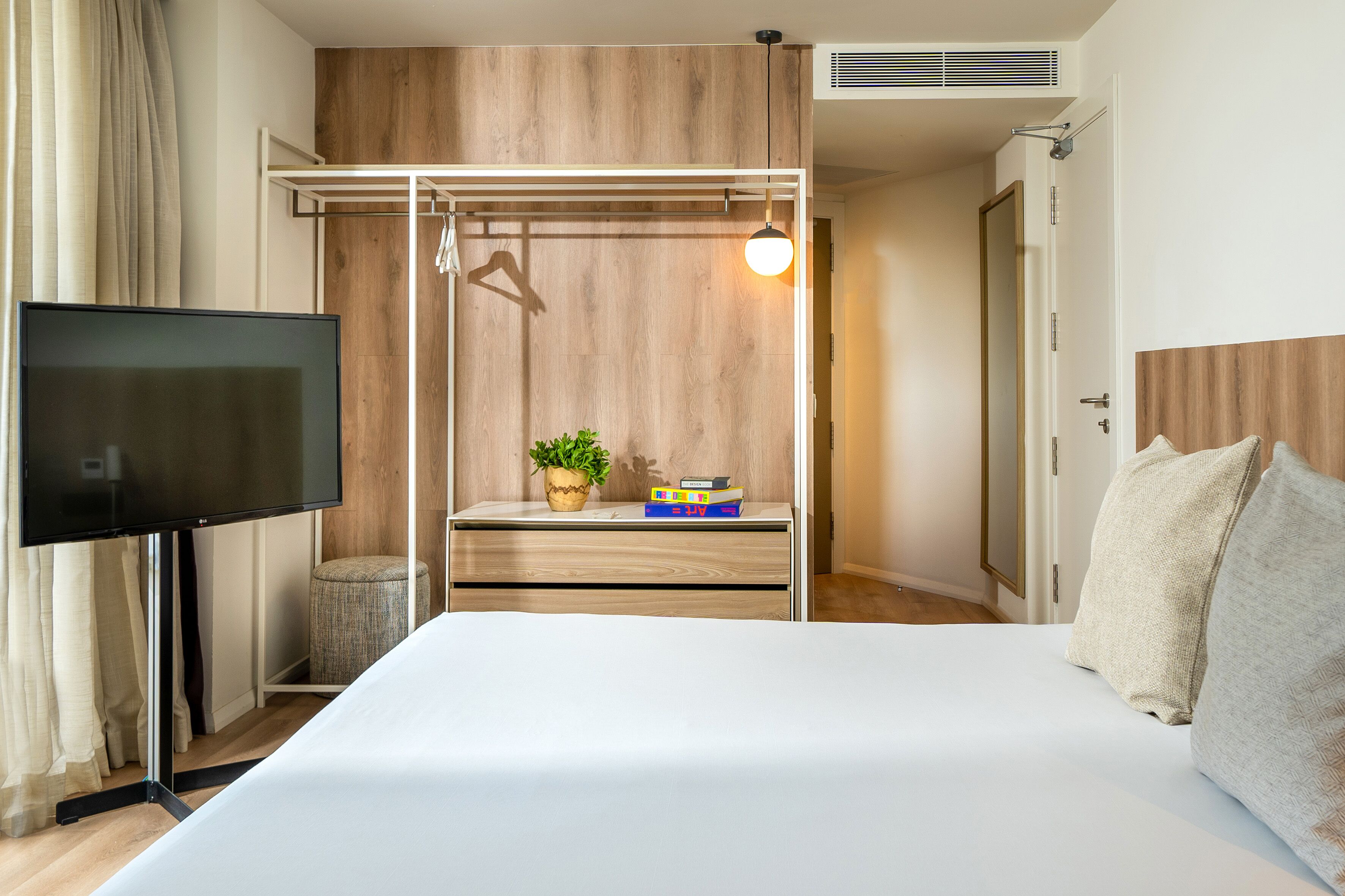 Ein Hotelzimmer mit einem Bett, einem Fernseher, einem Holzschrank, einem kleinen Tisch mit Pflanze und Büchern sowie einer Pendelleuchte.