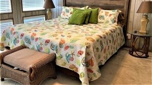 A bedroom with a wooden headboard, a floral bedspread, two green pillows, a wicker chair, and two bedside lamps. - Three Bedroom, Two Bath. Off Water Home. Short Distance to Public Pier. (Port Mansfield)