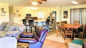 A living room with a sofa, a leather chair, a dining table with chairs, a kitchen area with a refrigerator, microwave, and bar stools. - Three Bedroom, Two Bath. Off Water Home. Short Distance to Public Pier. (Port Mansfield)