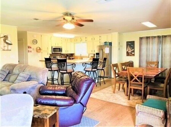 A living room with a sofa, a leather chair, a dining table with chairs, a kitchen area with a refrigerator, microwave, and bar stools.