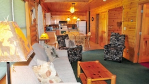 Interior acolhedor de uma cabana, com um sofá, duas poltronas estampadas, uma mesa de madeira e um espaço de jantar com cadeiras. Uma cozinha com eletrodomésticos e uma mesa de jantar é visível ao fundo.